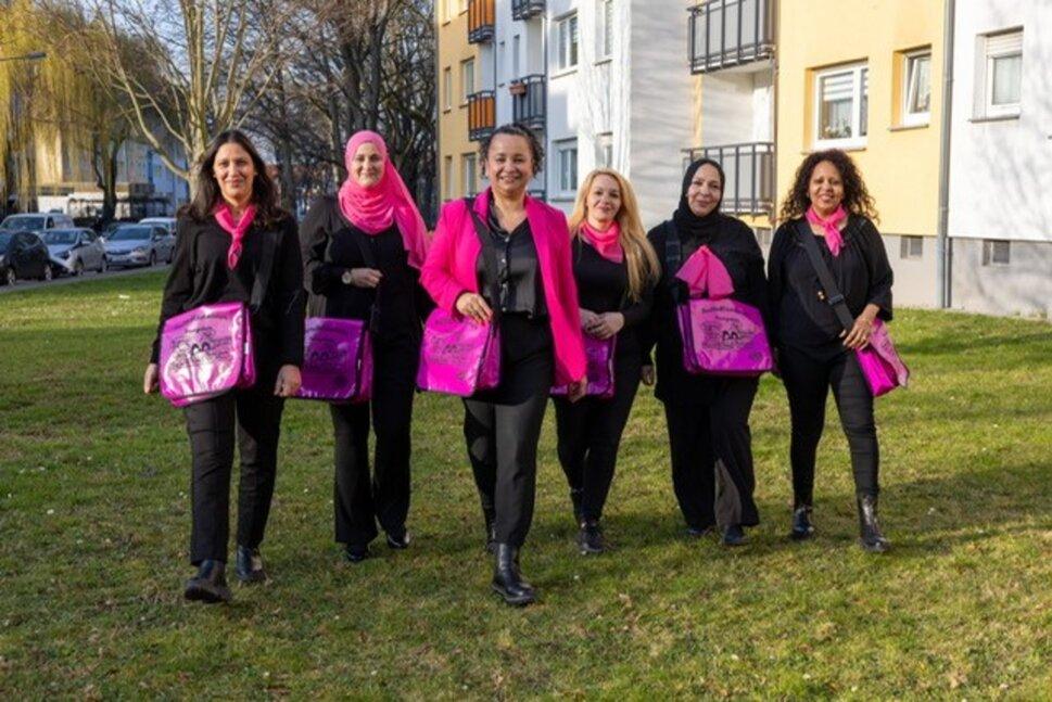 Sechs der Stadtteilflüsterinnen laufen lächelnd und in einer Reihe auf die Kamera zu. Sie tragen pinke und schwarze Kleidung und auf ihren Taschen steht ebenfalls Stadtteilflüsterinnen.