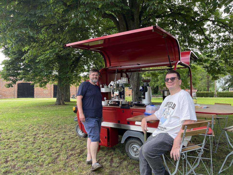 Das Café-Mobil steht aufgebaut auf einer Wiese. EIne Person steht an der Kaffeetheke. Eine Person sitzt auf einem Klappstuhl. Beide lächeln in die Kamera. Im Hintergrund ein Tisch.