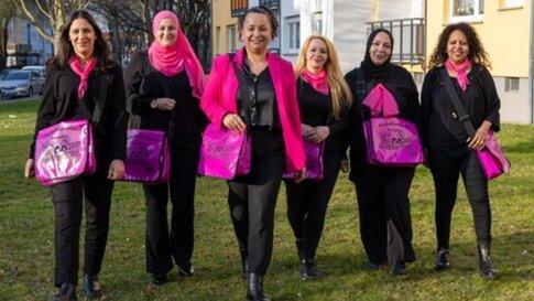 Sechs der Stadtteilflüsterinnen laufen lächelnd und in einer Reihe auf die Kamera zu. Sie tragen pinke und schwarze Kleidung und auf ihren Taschen steht ebenfalls Stadtteilflüsterinnen.