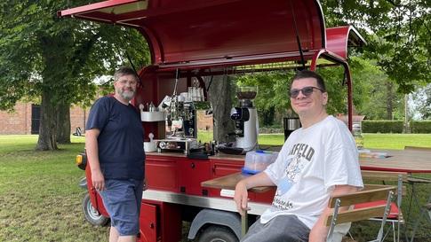 Das Café-Mobil steht aufgebaut auf einer Wiese. EIne Person steht an der Kaffeetheke. Eine Person sitzt auf einem Klappstuhl. Beide lächeln in die Kamera. Im Hintergrund ein Tisch.