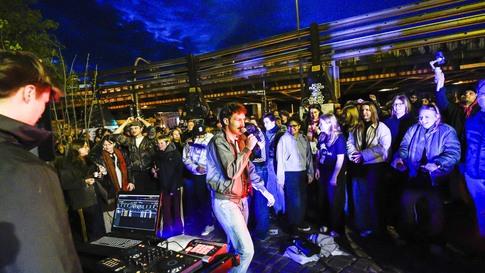 Eine große Gruppe an Menschen steht im Freien und sieht einer Person am Mikrofon zu, die singt. Es ist Abend.