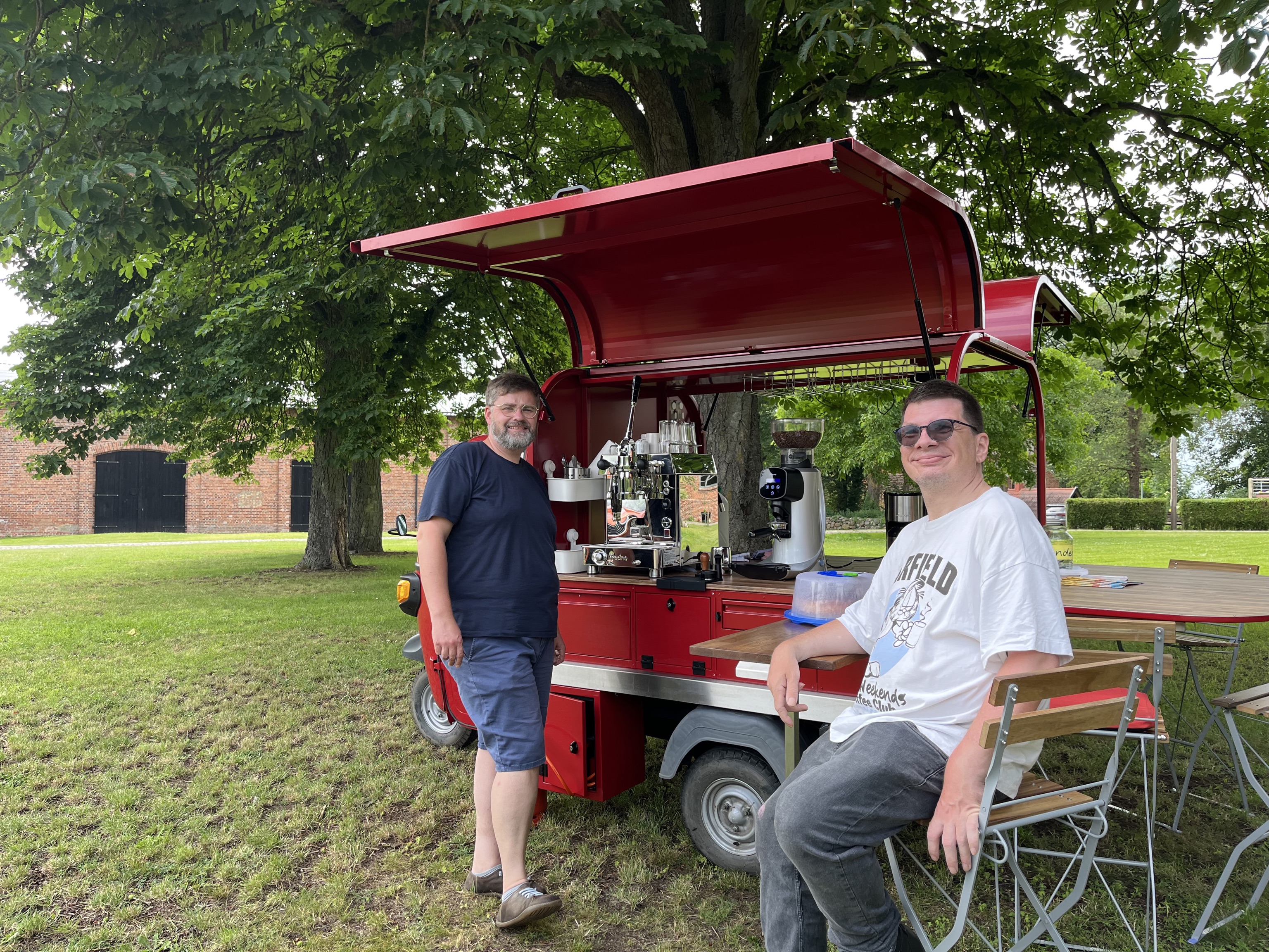 Das Café-Mobil steht aufgebaut auf einer Wiese. EIne Person steht an der Kaffeetheke. Eine Person sitzt auf einem Klappstuhl. Beide lächeln in die Kamera. Im Hintergrund ein Tisch.